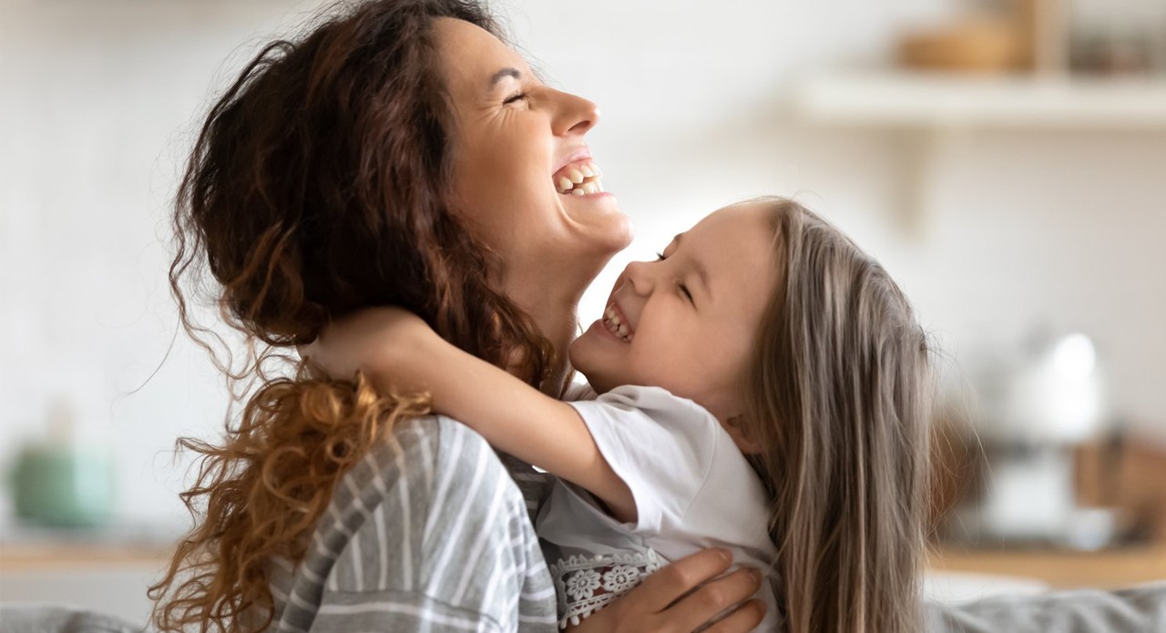 Adult with curly hair and young child embracing and laughing together in bright home interior with soft focus background.