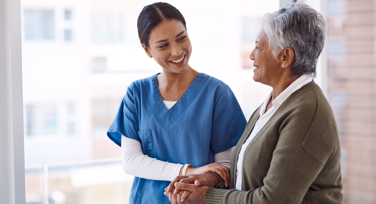 A healthcare worker in blue scrubs smiling warmly whilst holding the hands of an elderly woman with silver hair and a grey cardigan, both standing by a bright window.