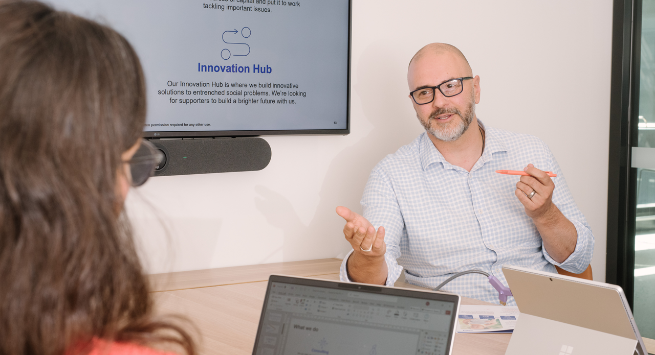 Person in checkered shirt presenting about Innovation Hub in meeting room, gesturing while explaining to colleague with laptop, screen displays company's social innovation mission.