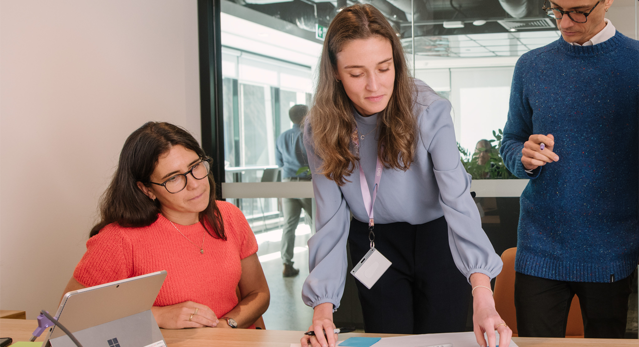 Three people collaborating in modern office, with person in coral sweater seated at laptop while person in light blue blouse with ID badge reviews documents, blue-sweater-wearing person partially visible to right.
