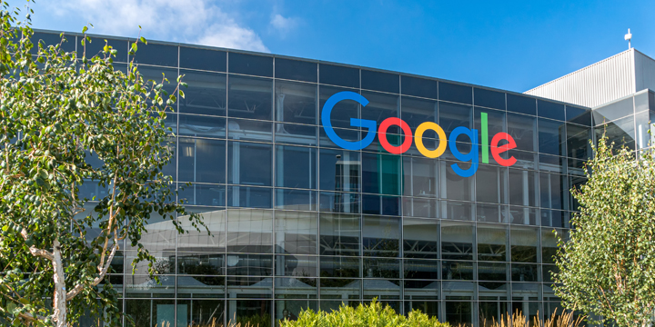 Google headquarters building with large colorful Google logo displayed on glass facade, surrounded by landscaped trees and plants under blue sky.