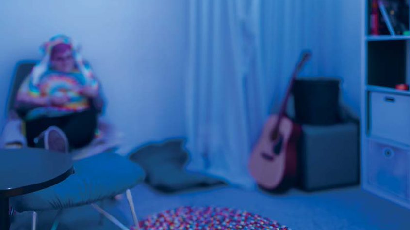 A bedroom with blue-tinted lighting showing a person sitting in a chair wearing a colorful patterned shirt. The room contains a bed with patterned bedding, an acoustic guitar leaning against furniture, bookshelves along the wall, and white curtains. The overall atmosphere has a moody, evening ambiance due to the blue lighting.