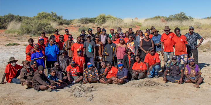 Large group of approximately 50-60 First Nation community members gathered outdoors in an landscape with red sand and sparse vegetation. Many wear bright orange shirts, arranged in multiple rows for a group photo.