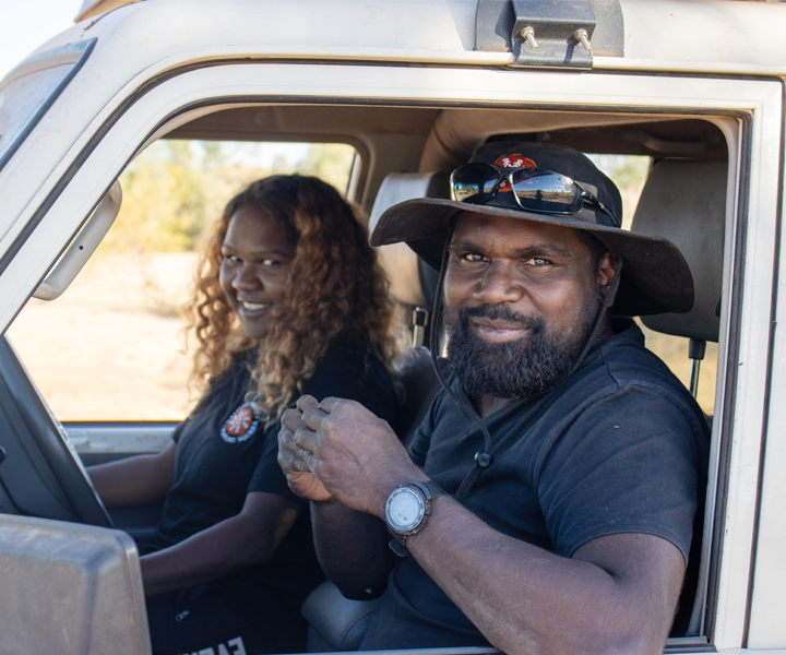 Two First Nation rangers in black uniforms sitting in a white vehicle, smiling at the camera.
