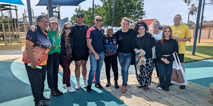 Group of eleven people posing together at a playground on a sunny day, with play equipment and trees visible in the background.