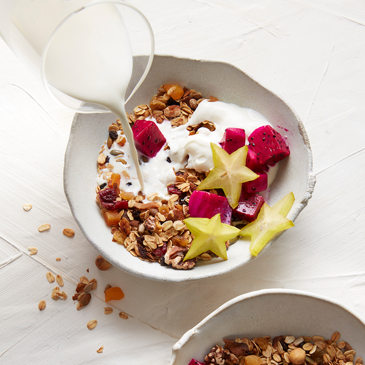 Bowl of granola topped with fresh fruit and yoghurt, with yoghurt being poured from a jug above the bowl.